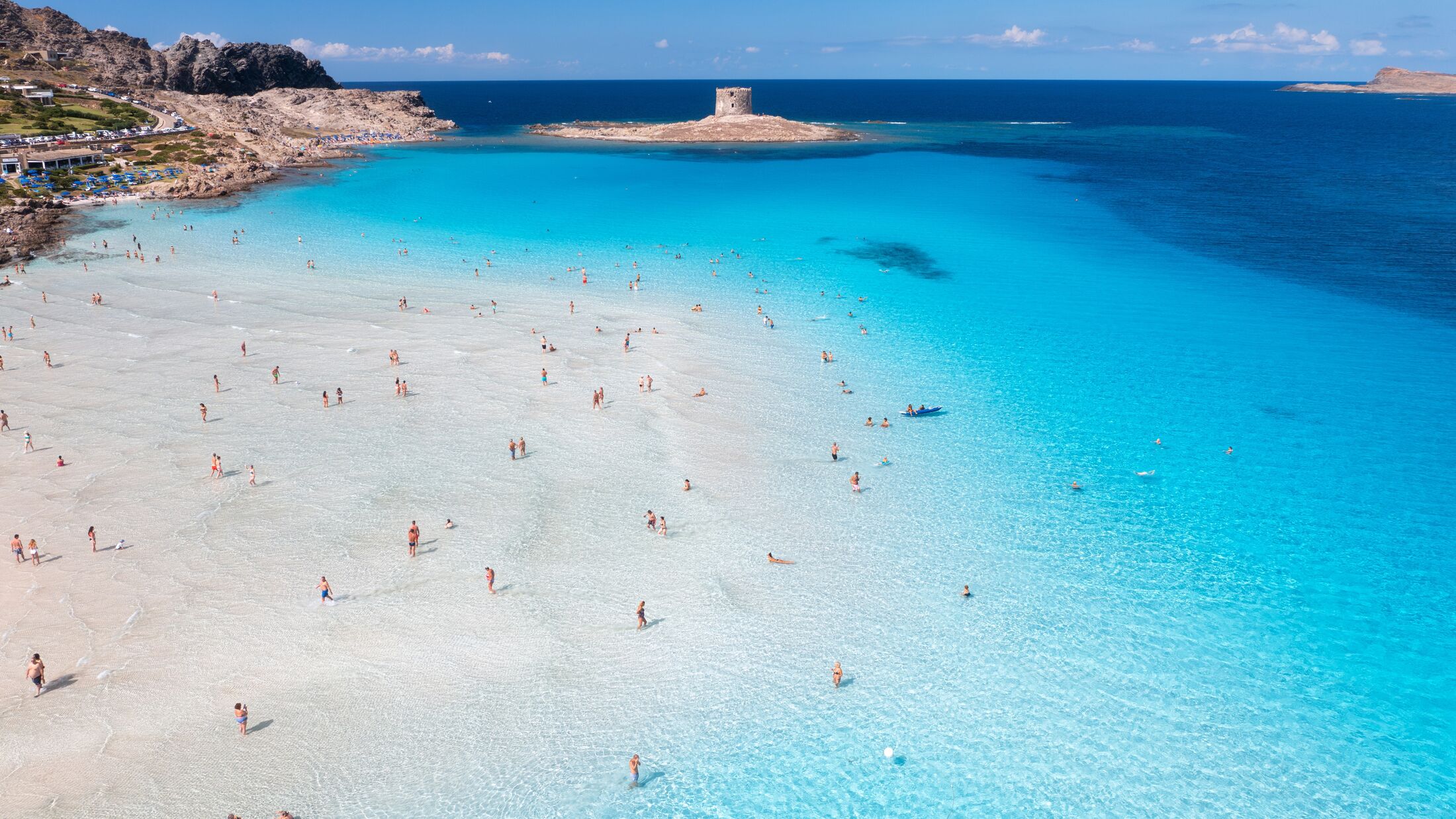 Aerial view of famous La Pelosa beach at sunny summer day. Stintino, Sardinia island, Italy. Top view of sandy beach, swimming people, clear blue sea, old tower and sky with clouds. Tropical seascape