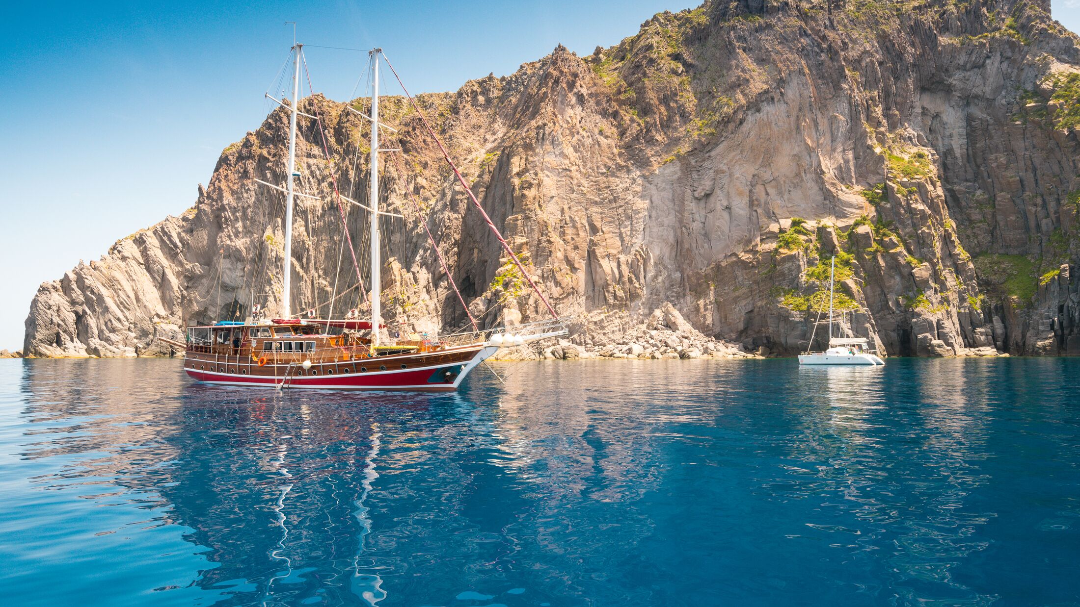 Wooden Classic Sailing yacht anchored in a deserted island in the Eolian Islands of the Mediterranean, with impressive cliffs and very blue sky and waters