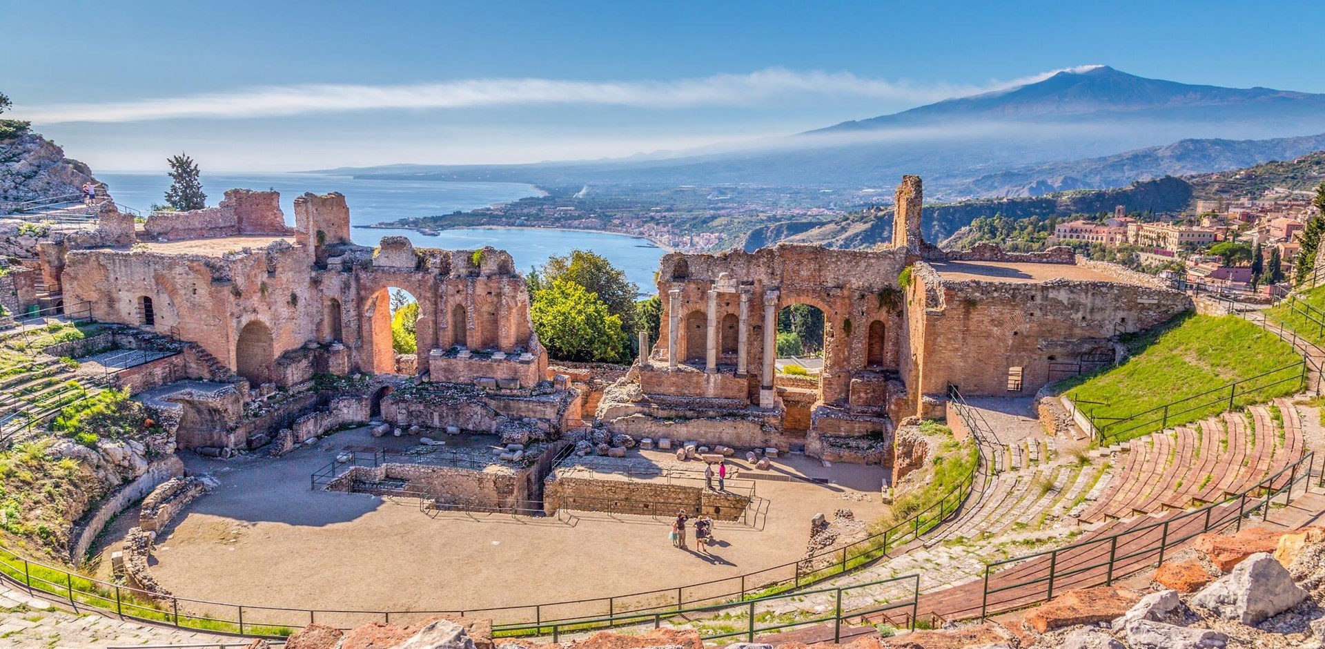 Ruins of the Ancient Greek Theater in Taormina, Sicily with the double smoke tail of the Etna extending over the the Giardini-Naxos bay of the Ionian Sea in the morning sun shine.