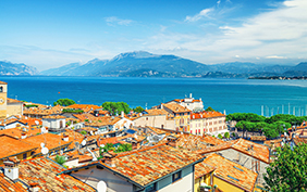Aerial panoramic view of Desenzano del Garda town with red tiled roof buildings, Garda Lake water surface, Monte Baldo mountain range, Sirmione peninsula, blue sky background, Lombardy, Northern Italy